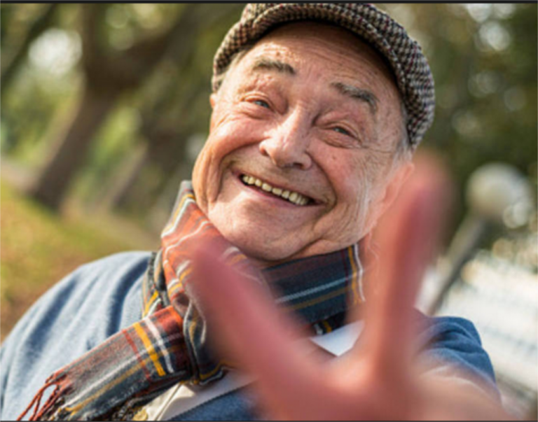 Senhor de boné com cachecol xadrez fazendo V de vitoria com os dedos e sorrindo